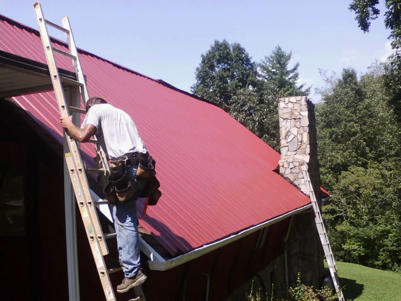 Roofer on a ladder beside a red metal roof and stone chimney — Soffit & Fascia Repair by Roofing Brothers in Cumberland City